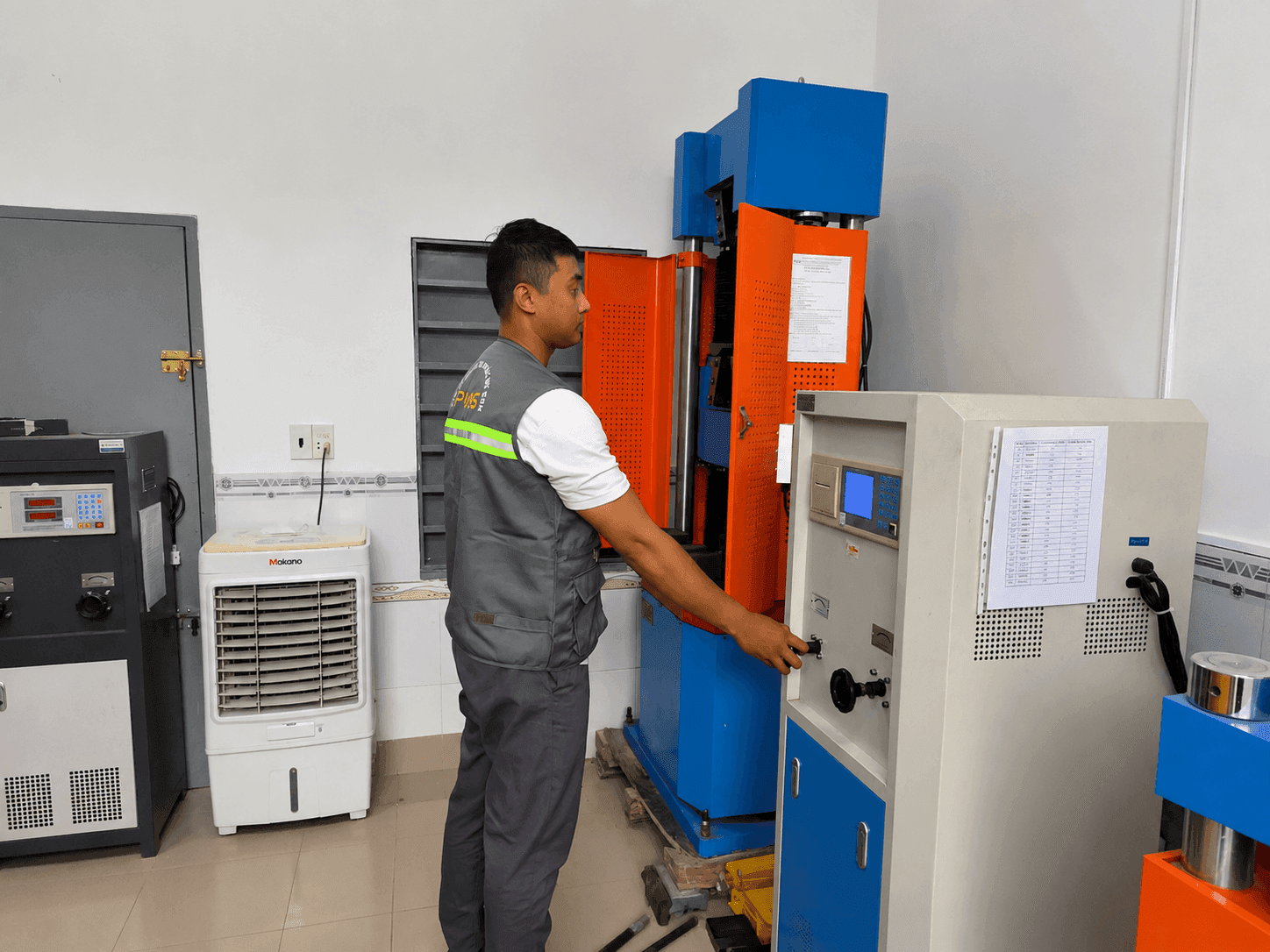A technician operates a large, blue and orange industrial testing machine in a lab.