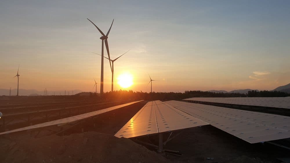 Solar panels reflect a sunset alongside wind turbines, symbolizing renewable energy generation.