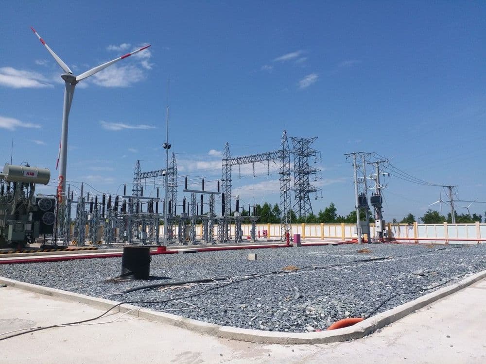 Electrical substation with power lines and a distant wind turbine under a blue sky.