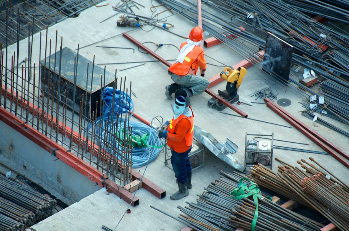 Construction workers in orange vests manage rebar and equipment on a concrete slab.