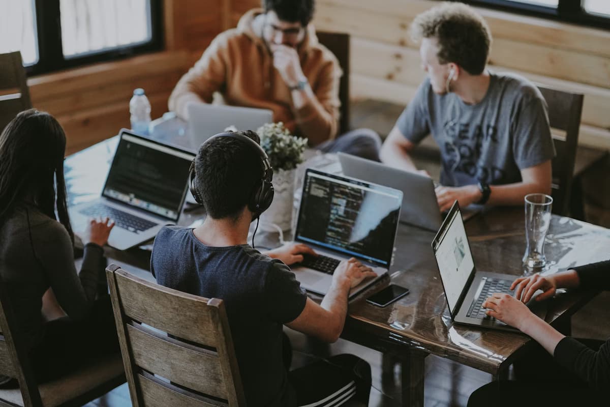 Group of young adults working collaboratively on laptops at a shared wooden table.