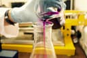 Gloved hand pouring vibrant purple chemical solution from beaker into an Erlenmeyer flask.