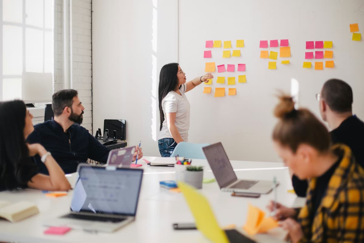 A woman points at colorful sticky notes on a whiteboard during a team brainstorming session.