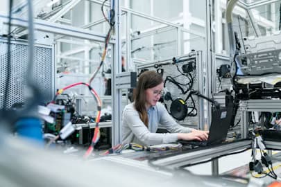 Female engineer uses a laptop amidst intricate wiring and machinery in a modern factory.
