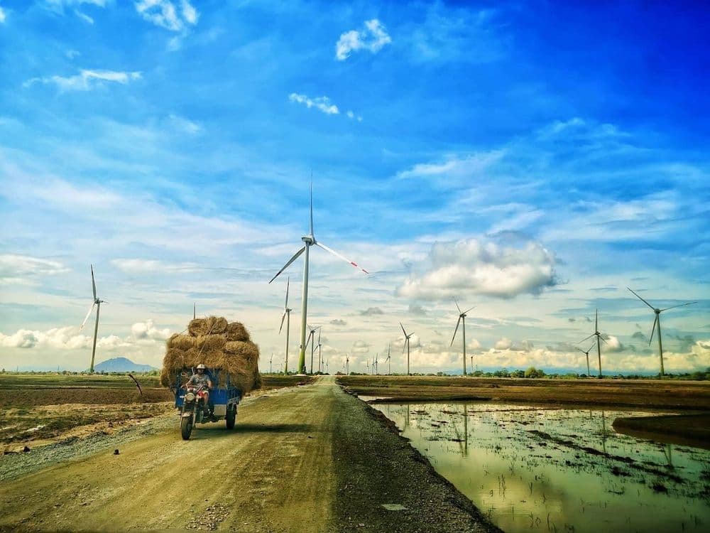 A hay-laden vehicle travels a rural road, flanked by wind turbines under a bright blue sky.