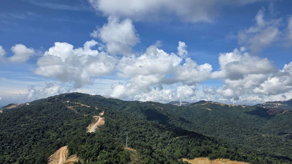 Lush green mountains under a blue sky with white clouds, showing cleared paths.