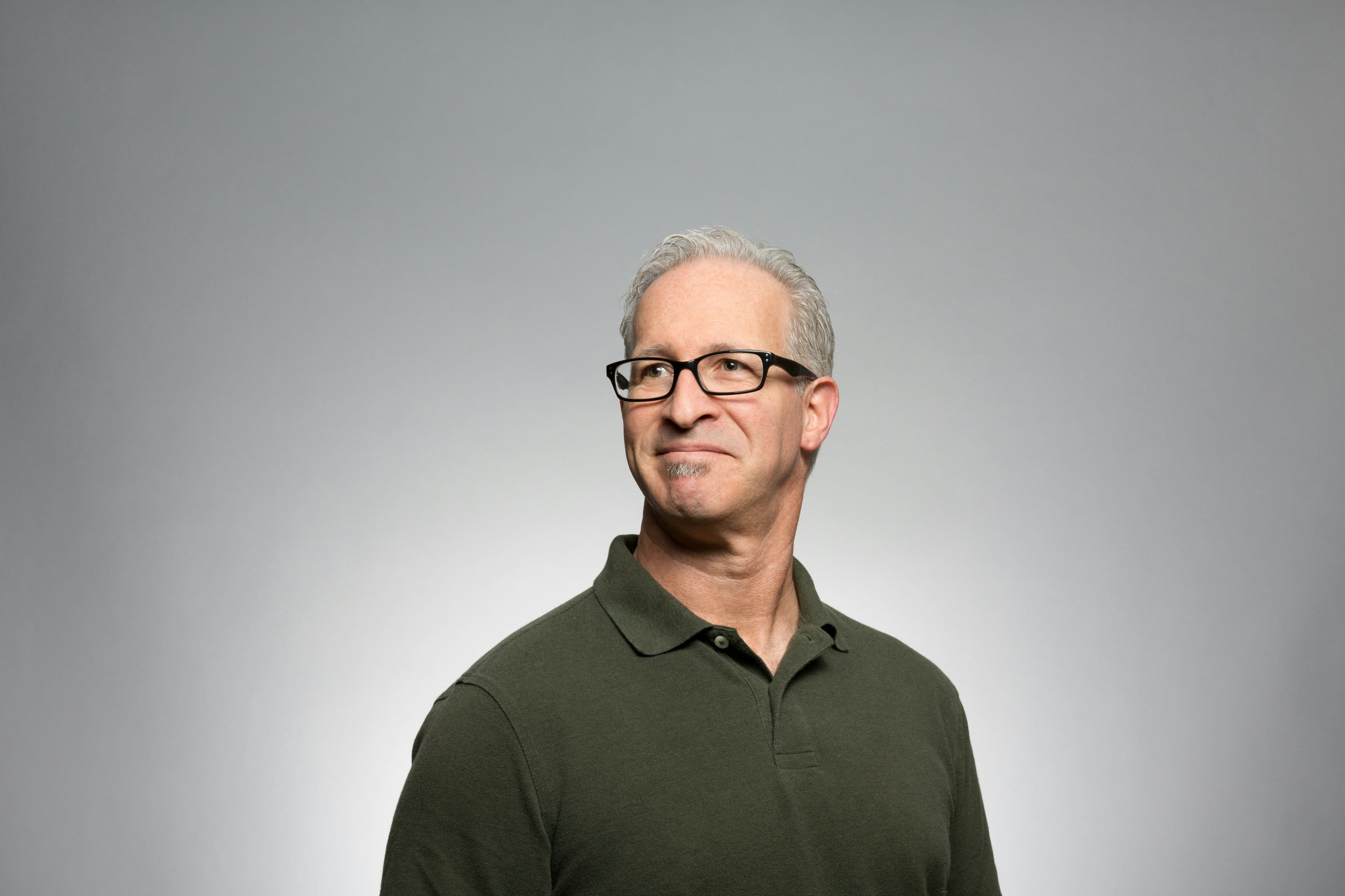 A man with grey hair and glasses, wearing a dark green polo shirt, looking left.