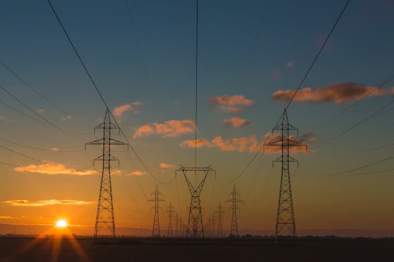 Electricity pylons are silhouetted against an orange and blue sunset with clouds.