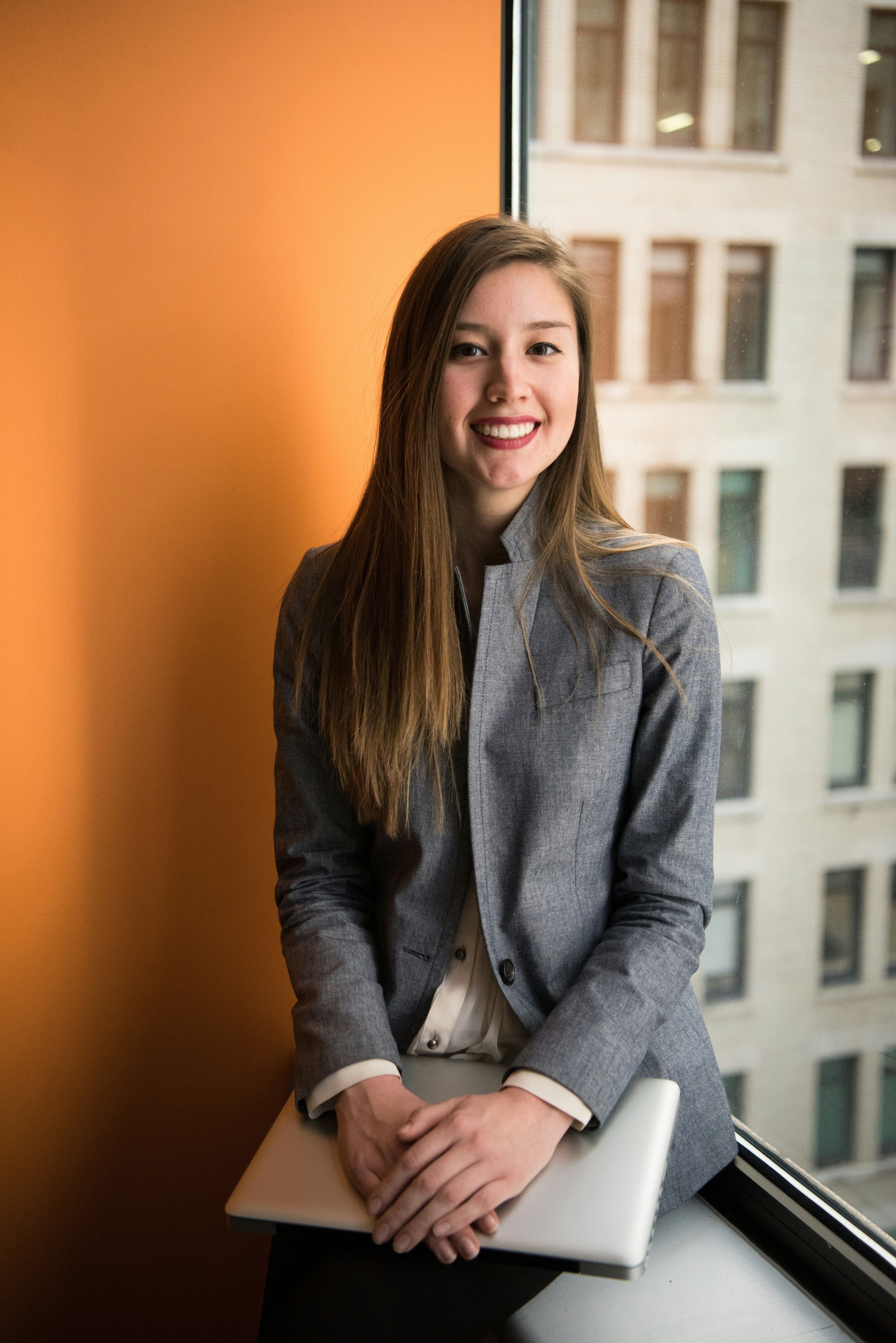 Smiling young woman in a gray blazer sits by a window holding a laptop.